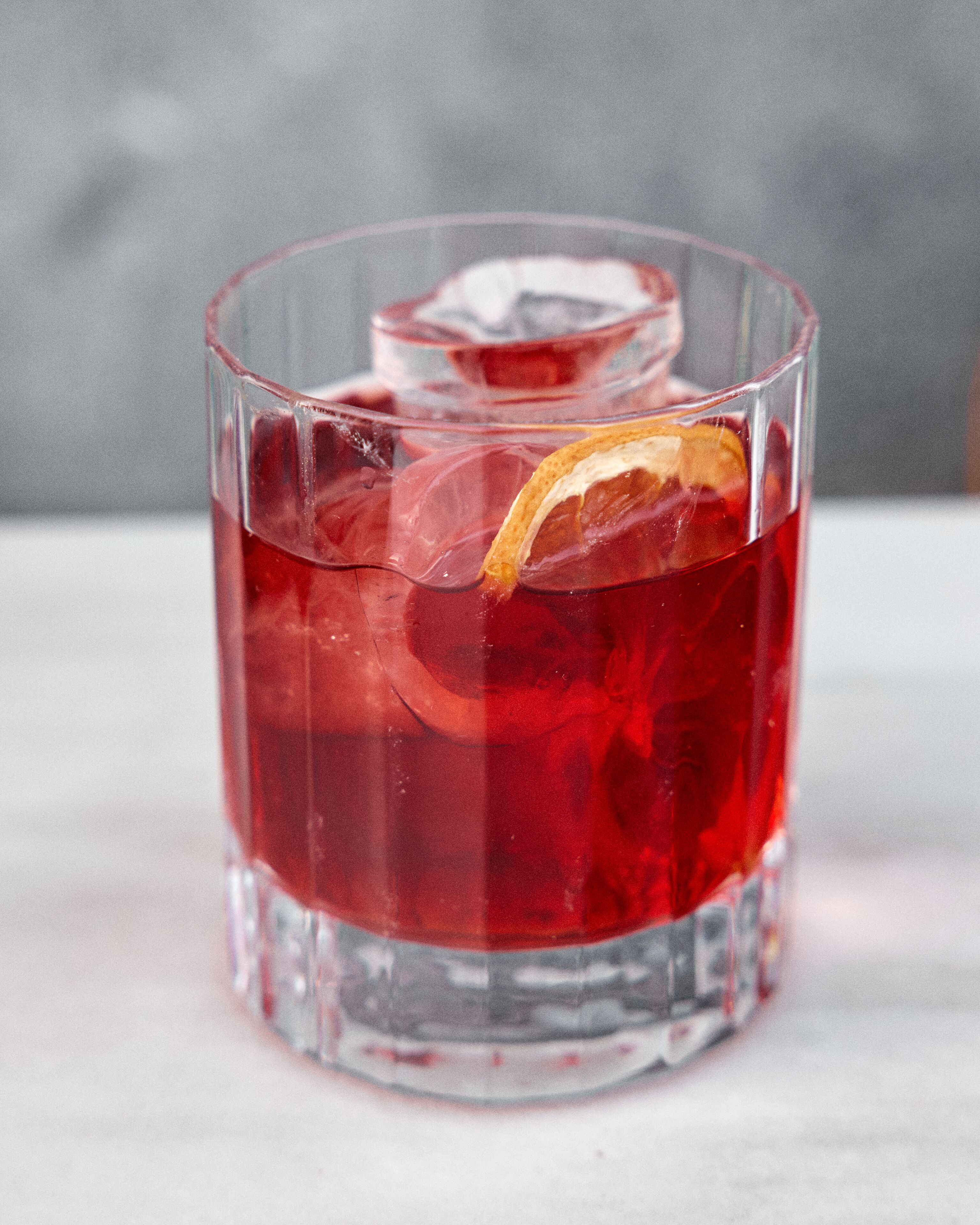Un vaso transparente con una bebida roja, cubitos de hielo y una rodaja de naranja.