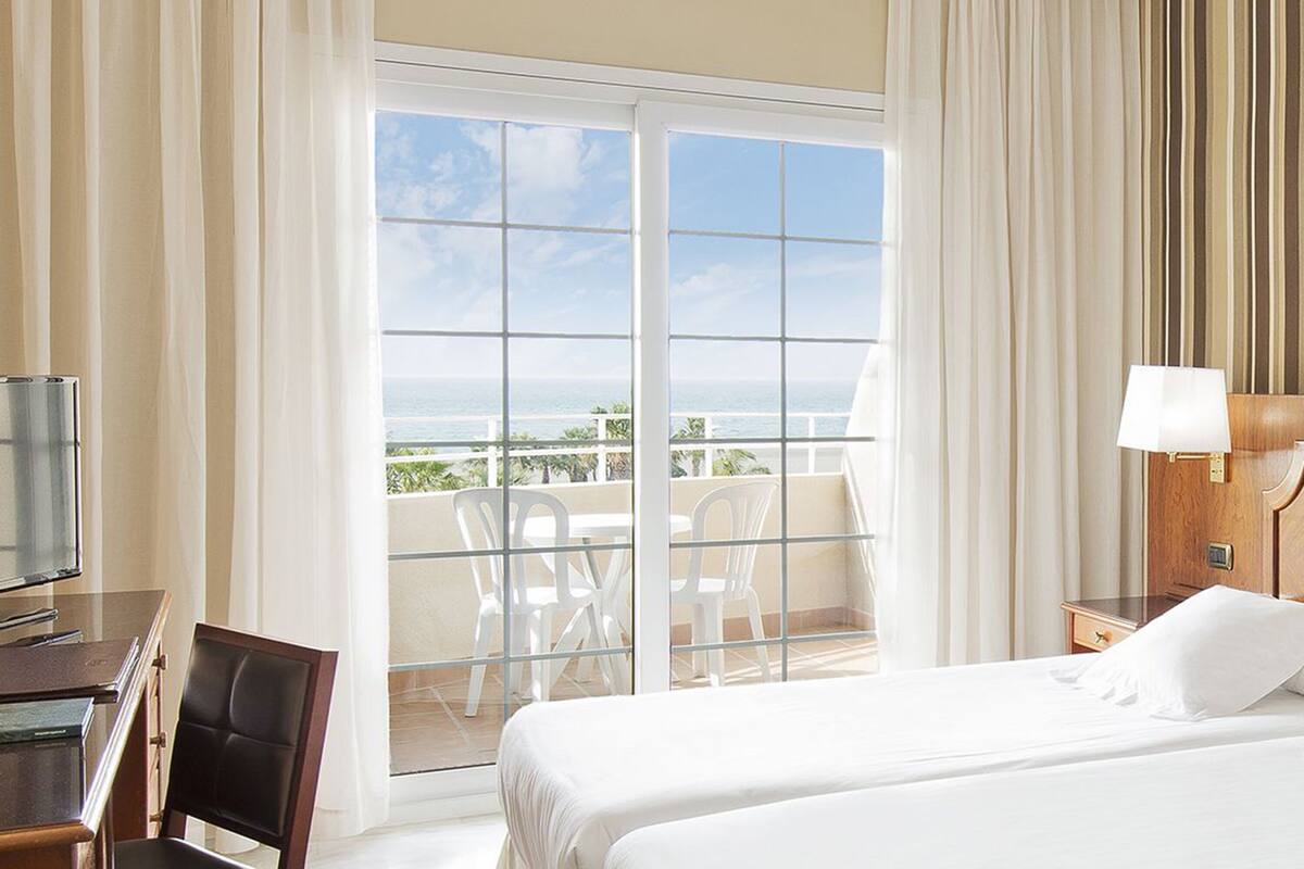 A hotel room with a bed, a desk, a TV, a balcony with a view of the ocean, and a window with curtains.