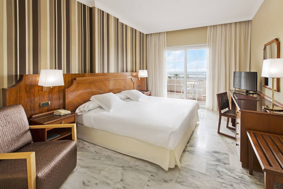 A hotel room with a large bed, a wooden headboard, a desk with a chair, a television, and a balcony with a view.