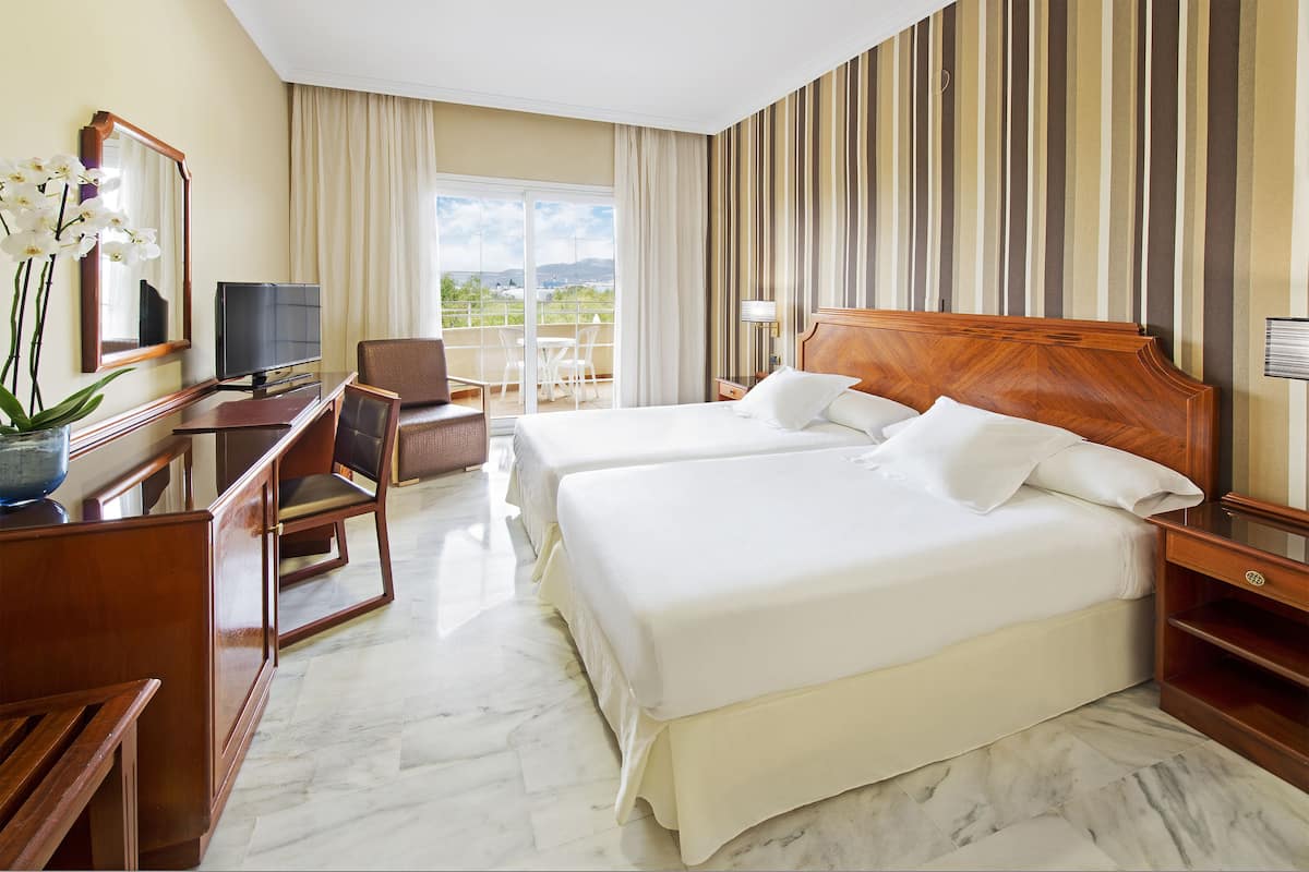 A hotel room with two beds, a desk with a TV, a chair, and a balcony with a view.