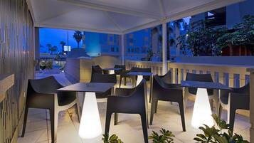 Un patio al aire libre con mesas y sillas, una zona cubierta y vistas a edificios y palmeras.