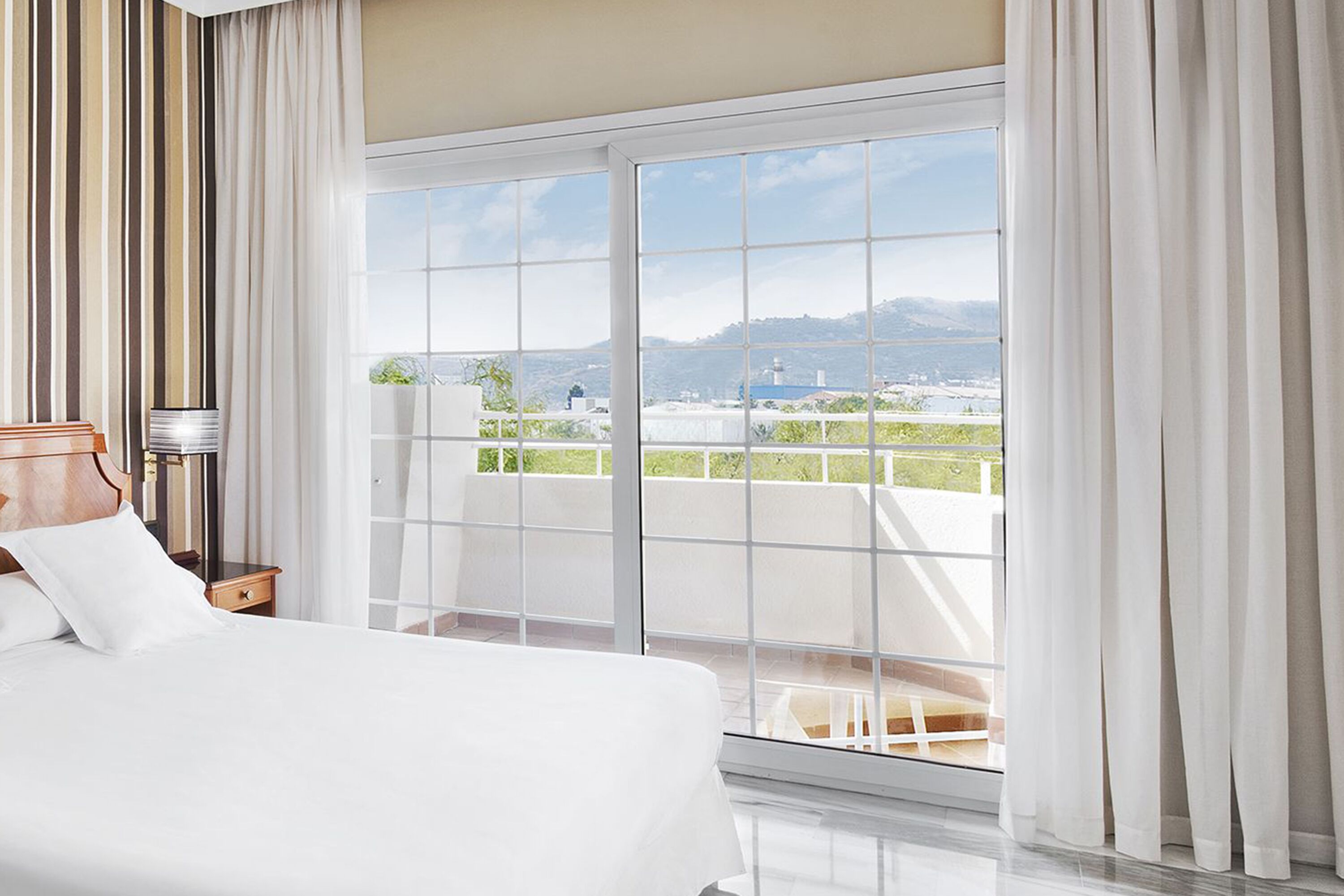 Een hotelkamer met een groot bed, een nachtkastje, een lamp en uitzicht op een stadsgezicht door middel van ramen van de vloer tot aan het plafond.