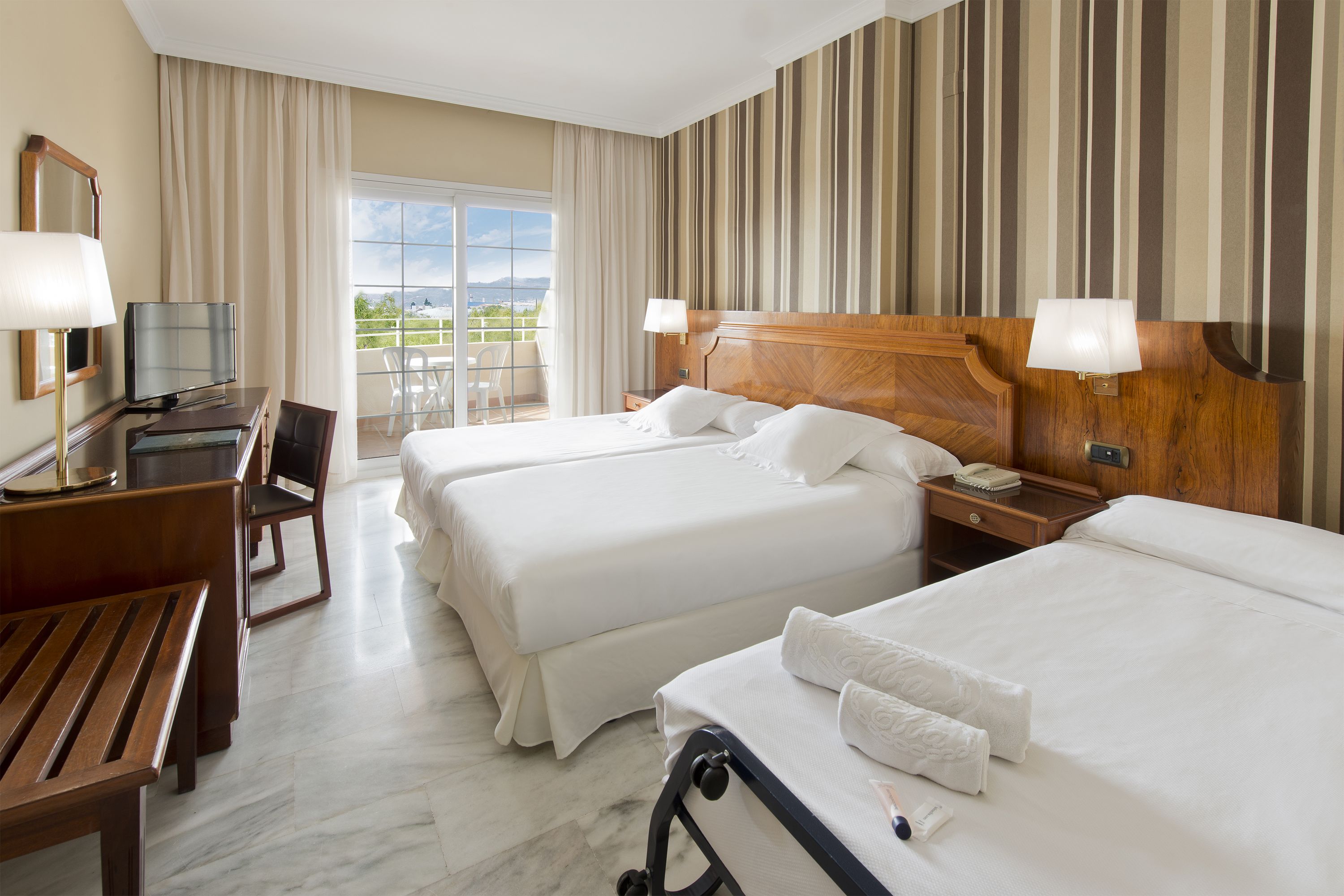 A hotel room with two beds, a desk with a TV, a chair, and a balcony with a view.