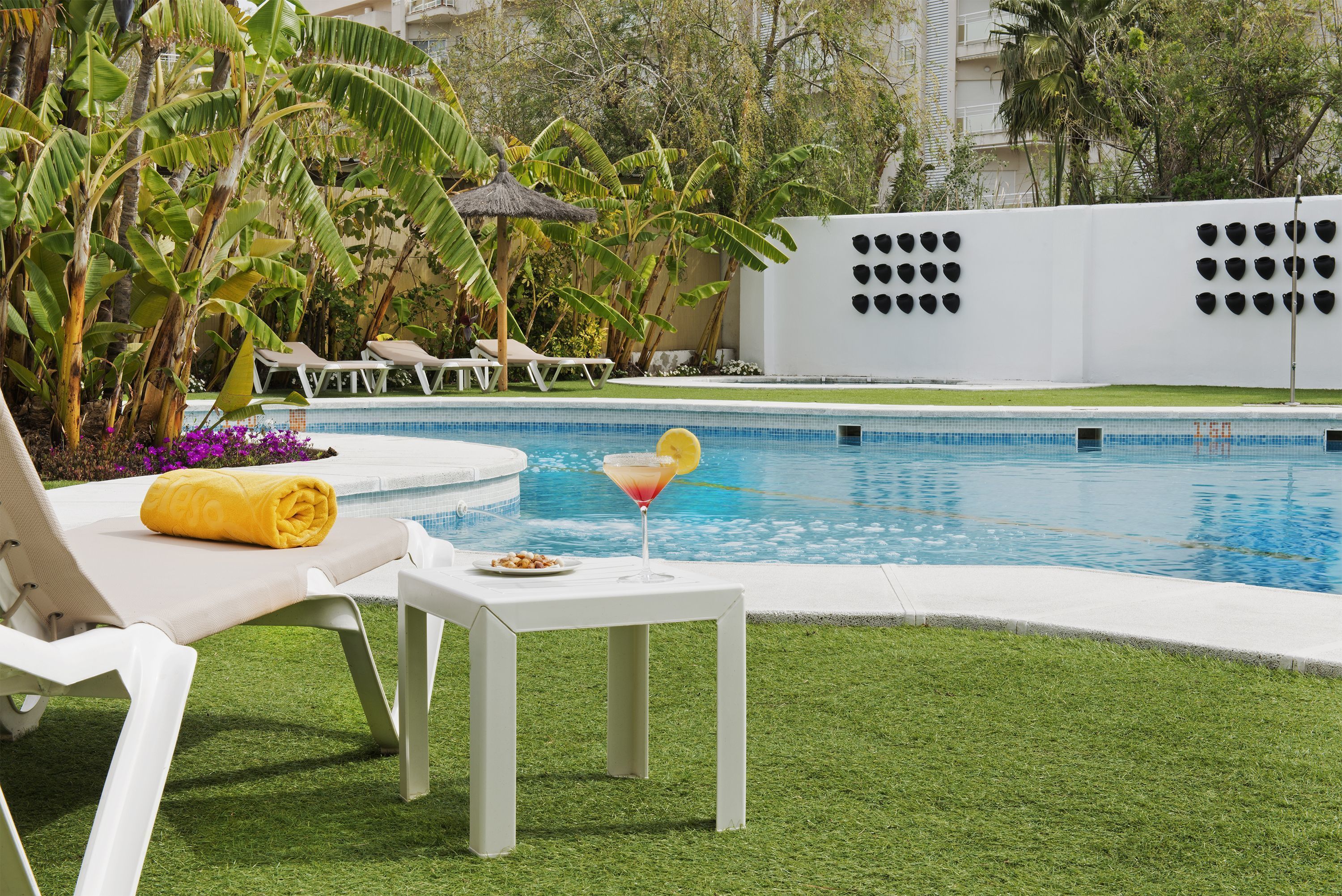 Un área de piscina con sillones de descanso, una mesa con una bebida y un jardín tropical.
