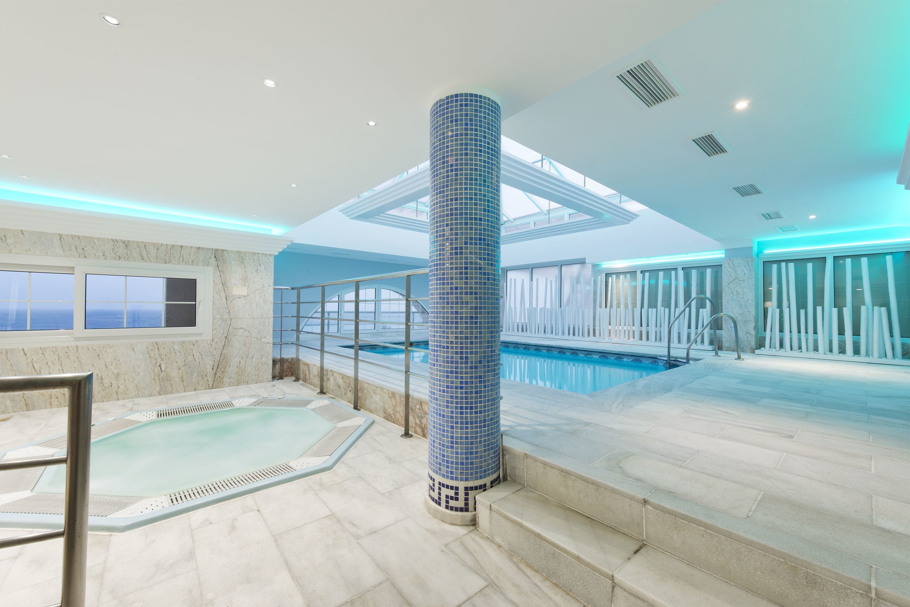 A modern indoor swimming pool with a tiled column, clear glass railings, and a view of the ocean through large windows.