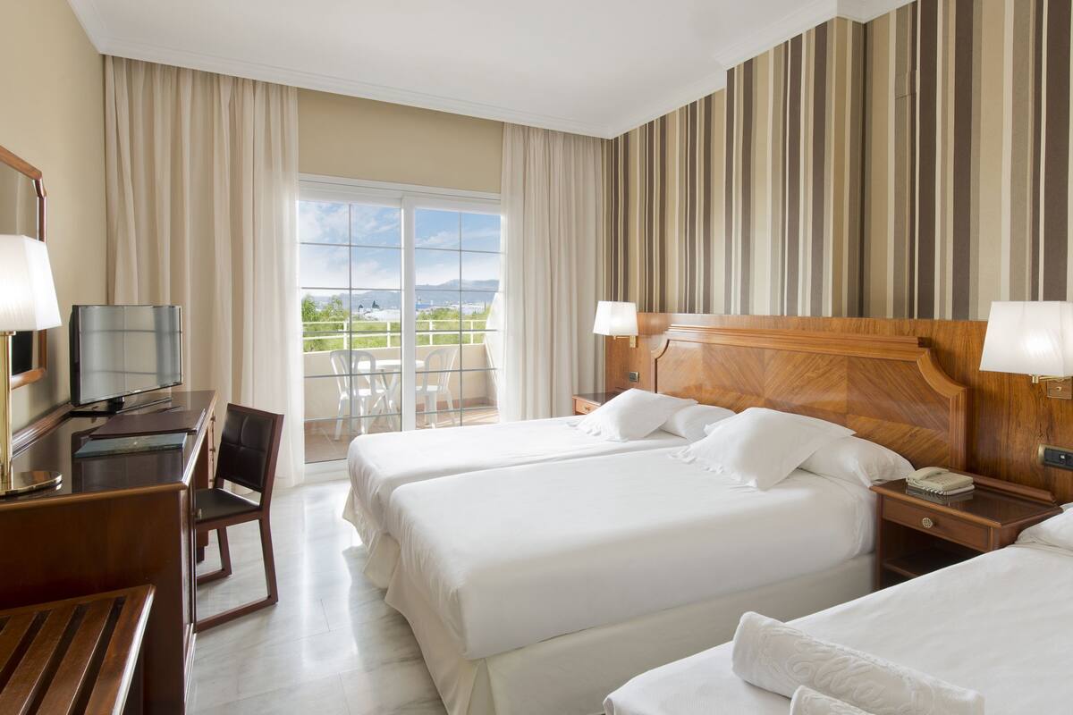 A hotel room with two beds, a desk with a television, a chair, and a balcony with a view.