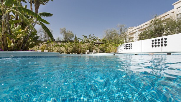 A clear blue pool with a tiled edge, surrounded by lush greenery and a white building with balconies. - Elba Motril Beach & Business Resort (Motril)