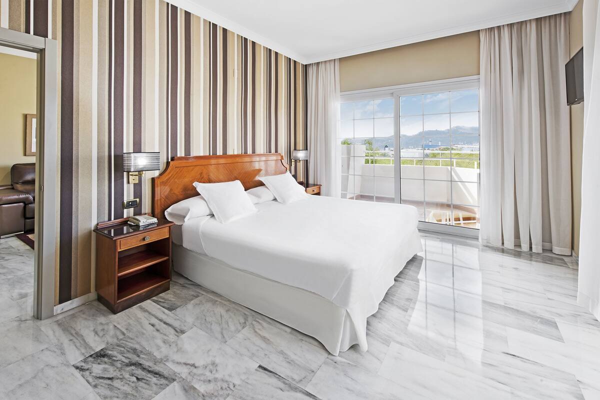 A hotel room with a large bed, a nightstand, a lamp, and a view of the outdoors.