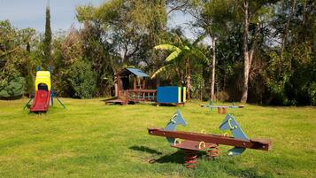 Kids can play on the slide, swing set, and seesaw in this outdoor playground surrounded by lush greenery.
