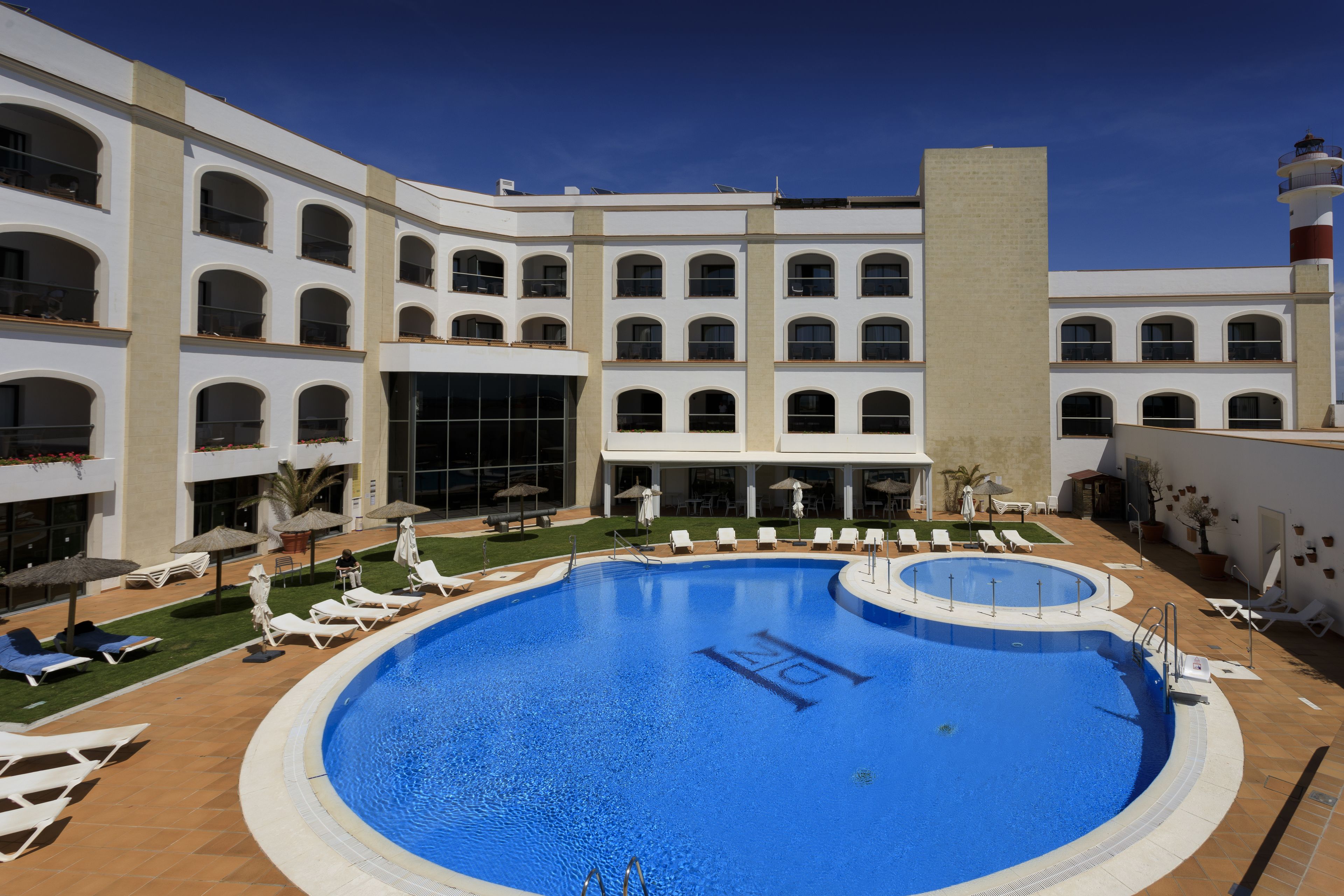 Uma grande piscina ao ar livre rodeada por espreguiçadeiras e guarda-sóis, com um edifício de vários andares ao fundo.