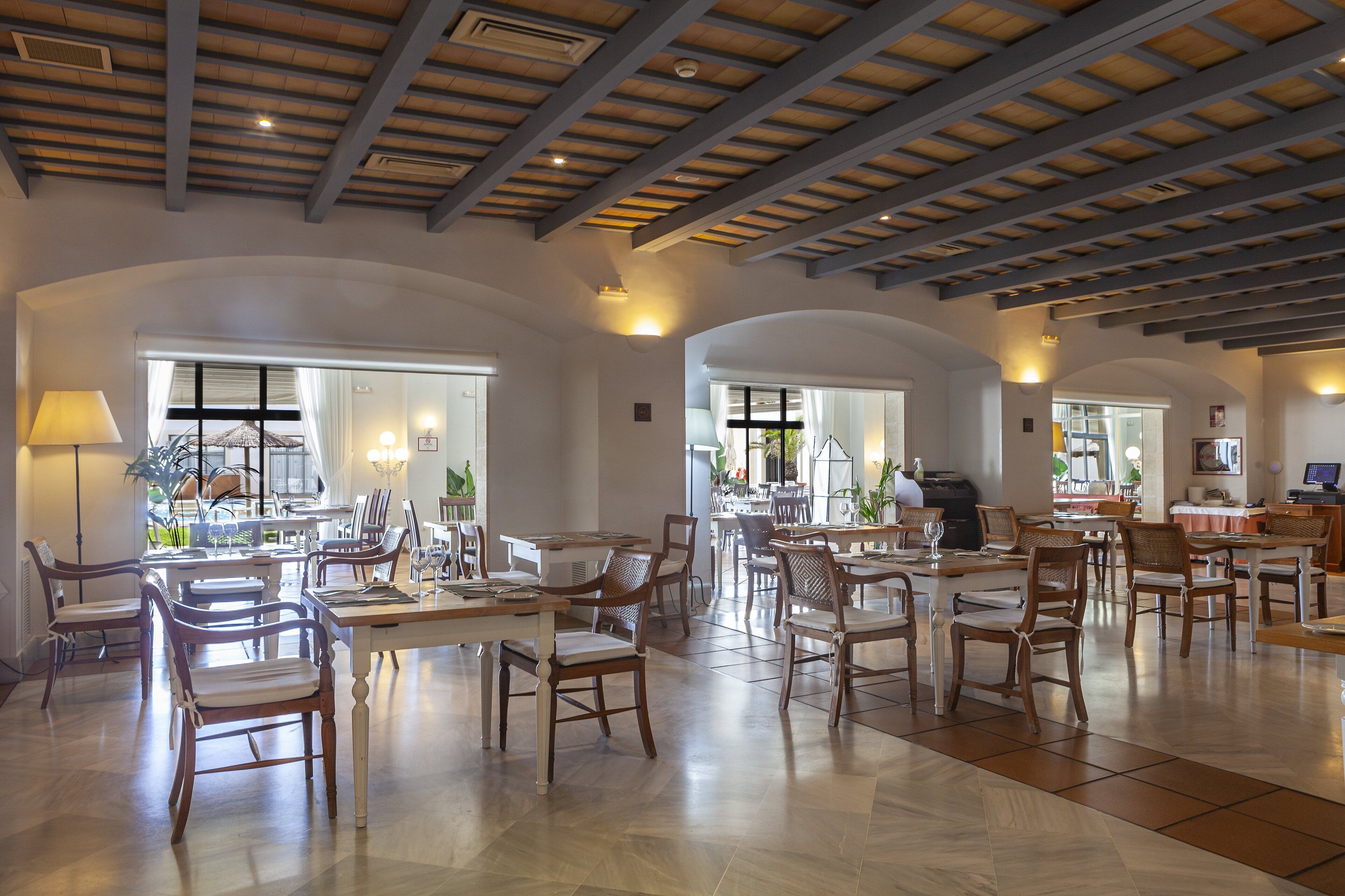 A spacious dining area with wooden tables and chairs, large windows, and a high ceiling with exposed beams.