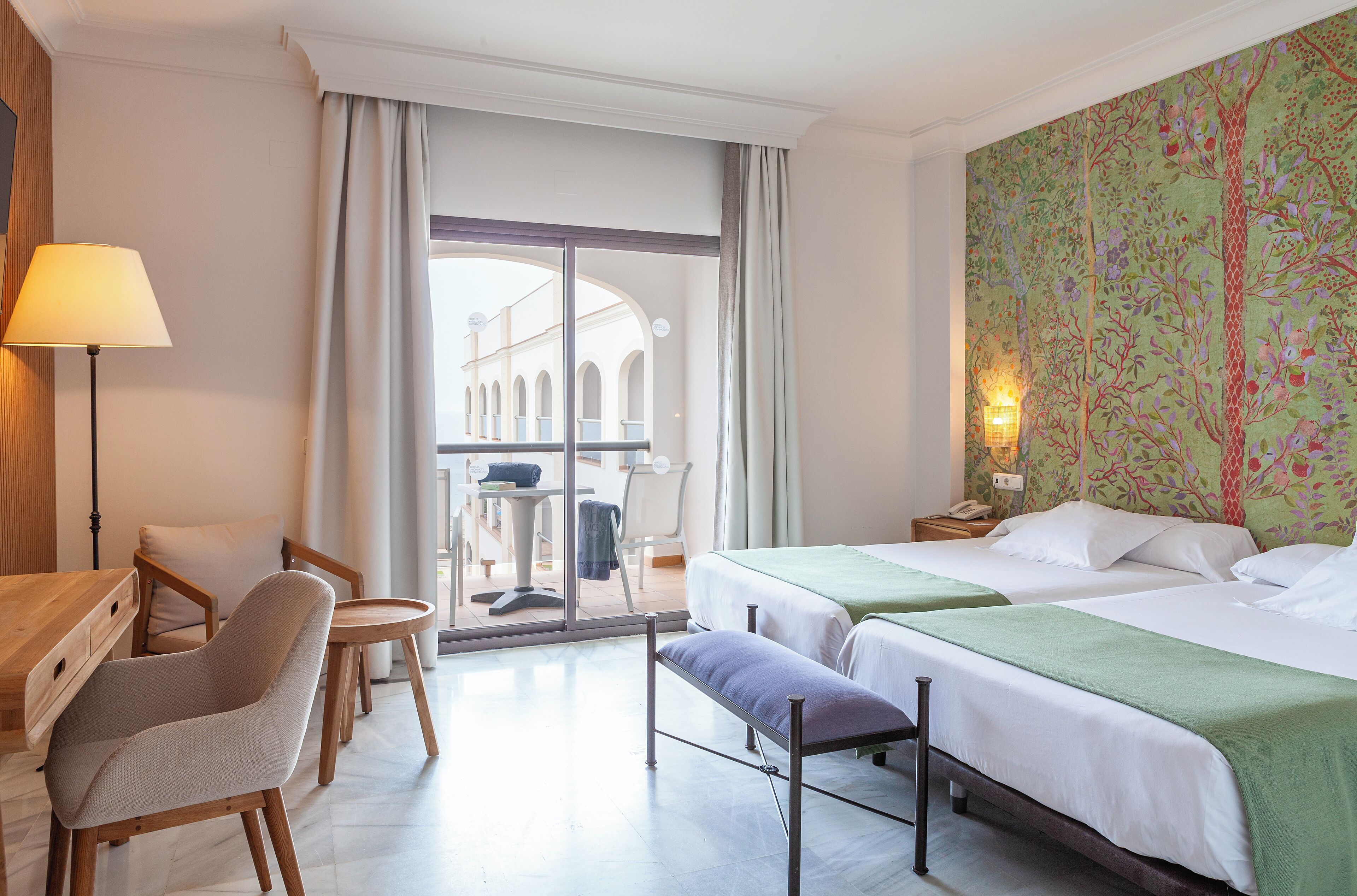 A hotel room with two beds, a desk, a chair, and a view of an outdoor area with a table and chairs.