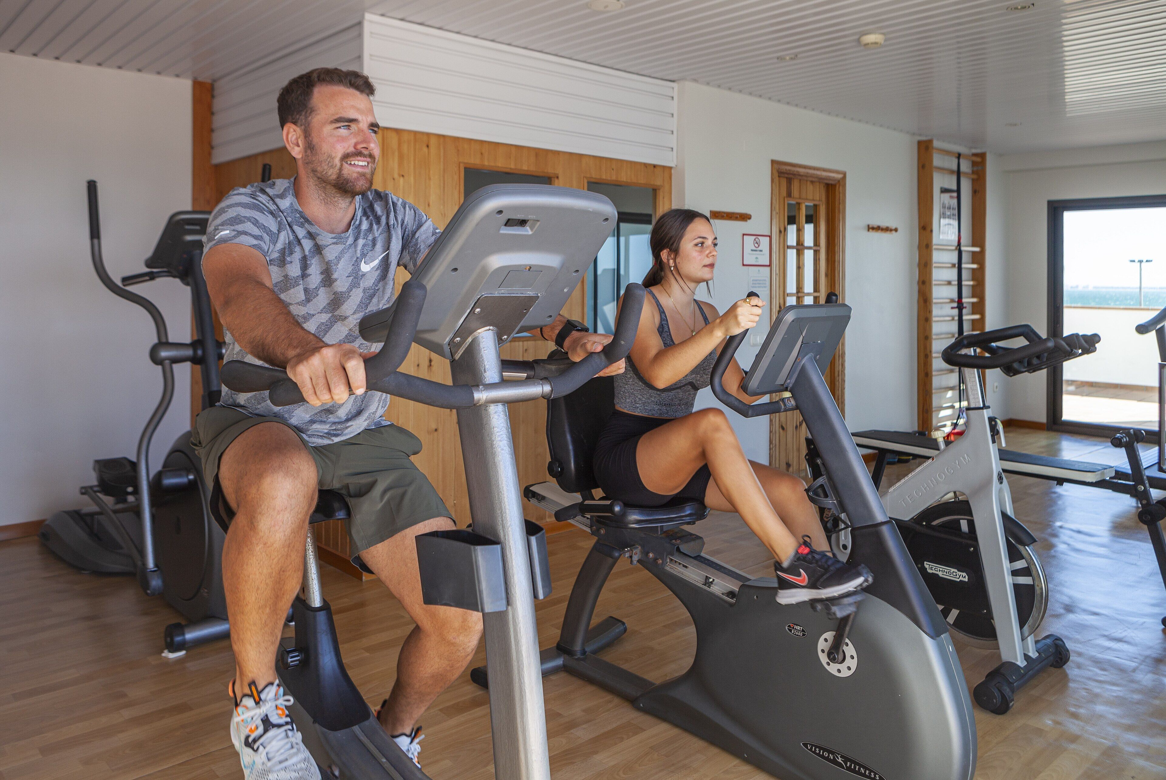 Two people exercising on stationary bikes in a gym with wooden flooring and a large window.