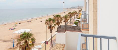 Vista de una playa desde un balcón con palmeras, un edificio blanco y cielo despejado.