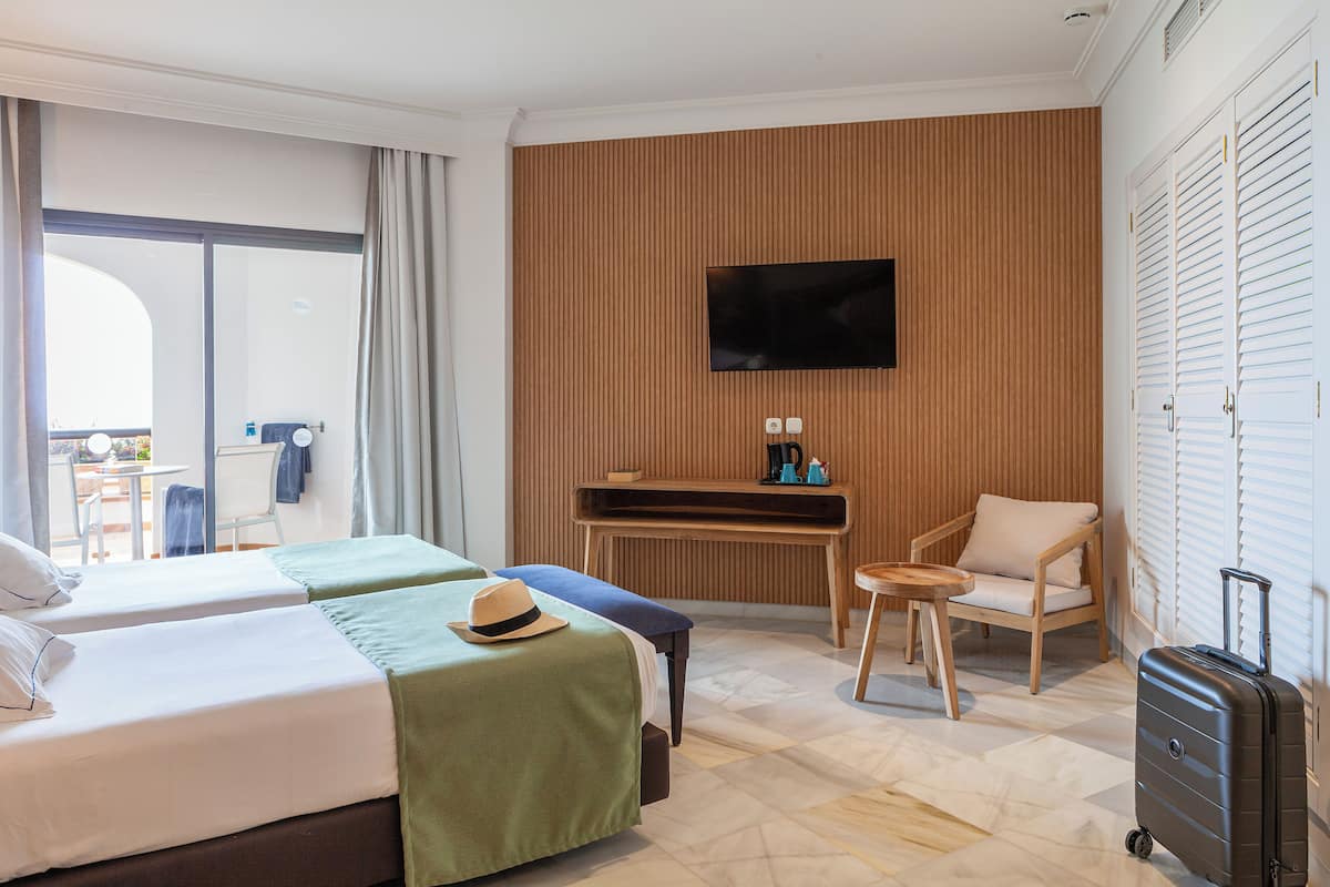 Habitación de hotel con dos camas, un televisor montado en la pared, un escritorio pequeño, una silla y una maleta.