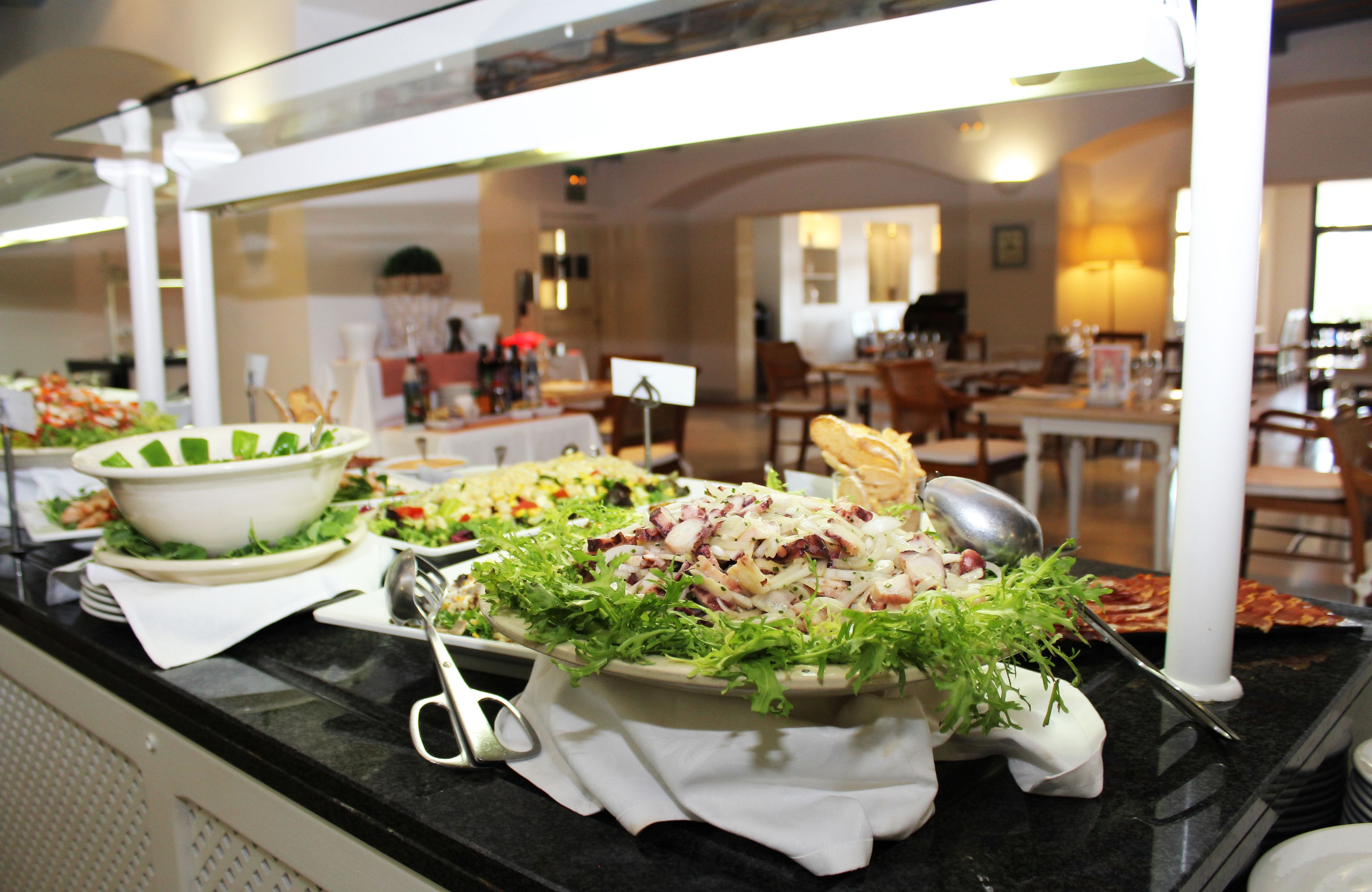 Een buffet met diverse gerechten, waaronder salades en een pastagerecht, in een goed verlicht eetgedeelte met tafels en stoelen.