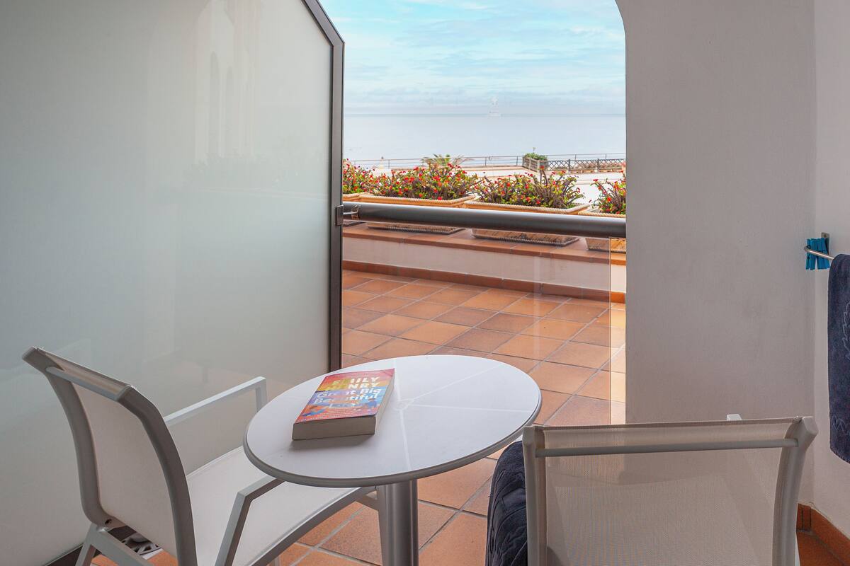 Un balcón con una mesa y sillas, un libro sobre la mesa, y vista al mar.