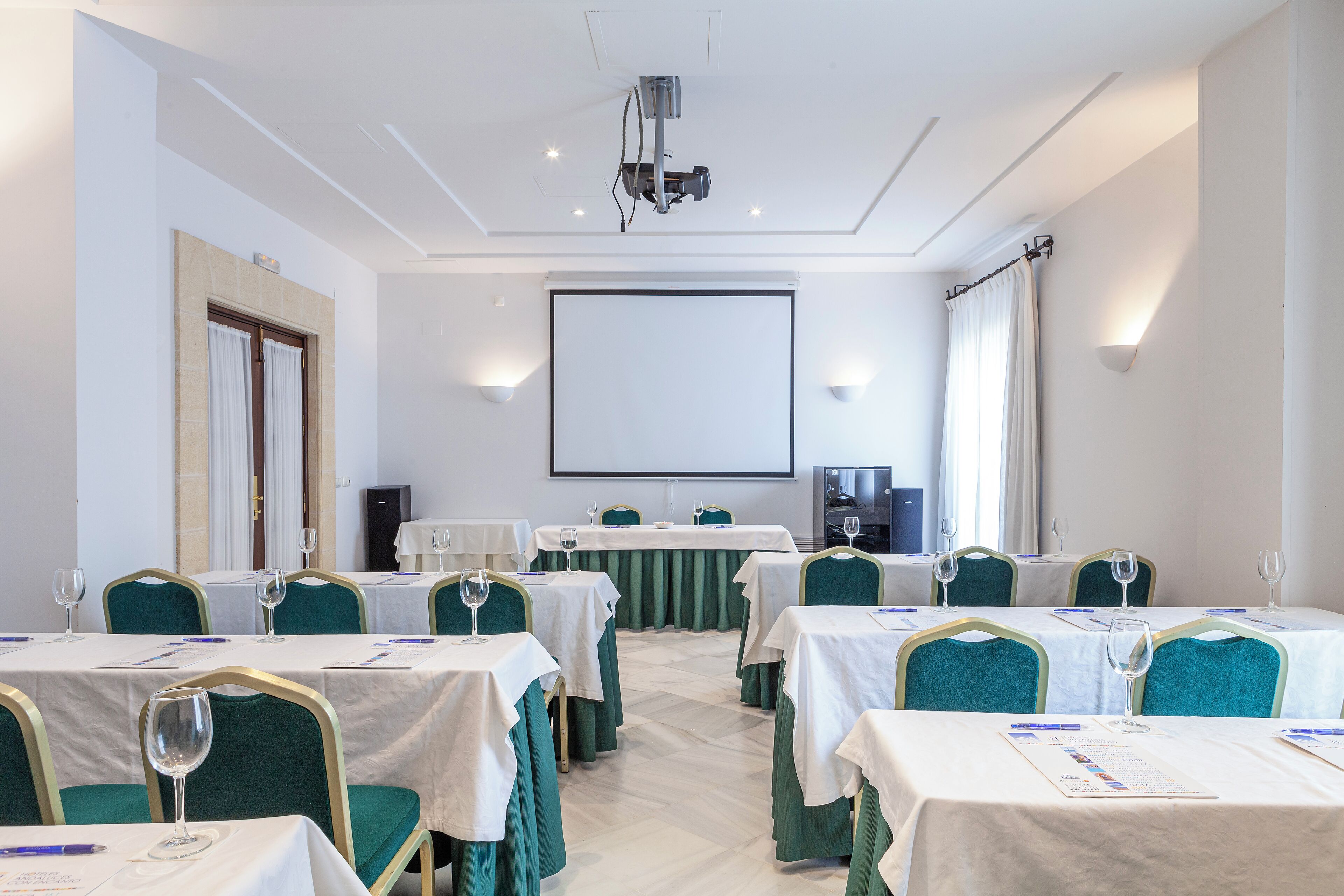Un salón de conferencias con mesas dispuestas para una reunión, un proyector y una pantalla.