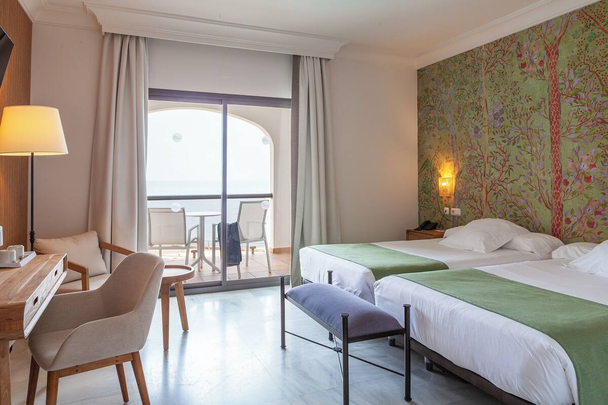Habitación de hotel con balcón, escritorio con silla, dos camas y una pared con estampado floral.