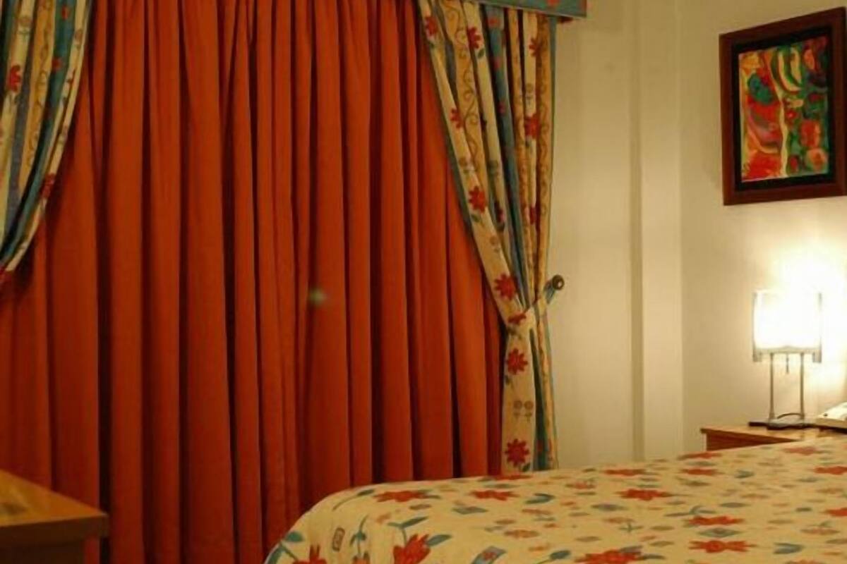 Ein Hotelzimmer mit Bett, Vorhängen, einem Bild an der Wand und einem Nachttisch mit Lampe.