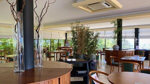 Un amplio comedor interior con mesas y sillas de madera, plantas grandes en macetas y vistas al verdor exterior.