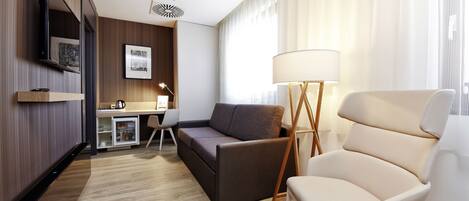Una habitación de hotel moderna con sofá, sillón, escritorio y televisor de pantalla plana montado en la pared.
