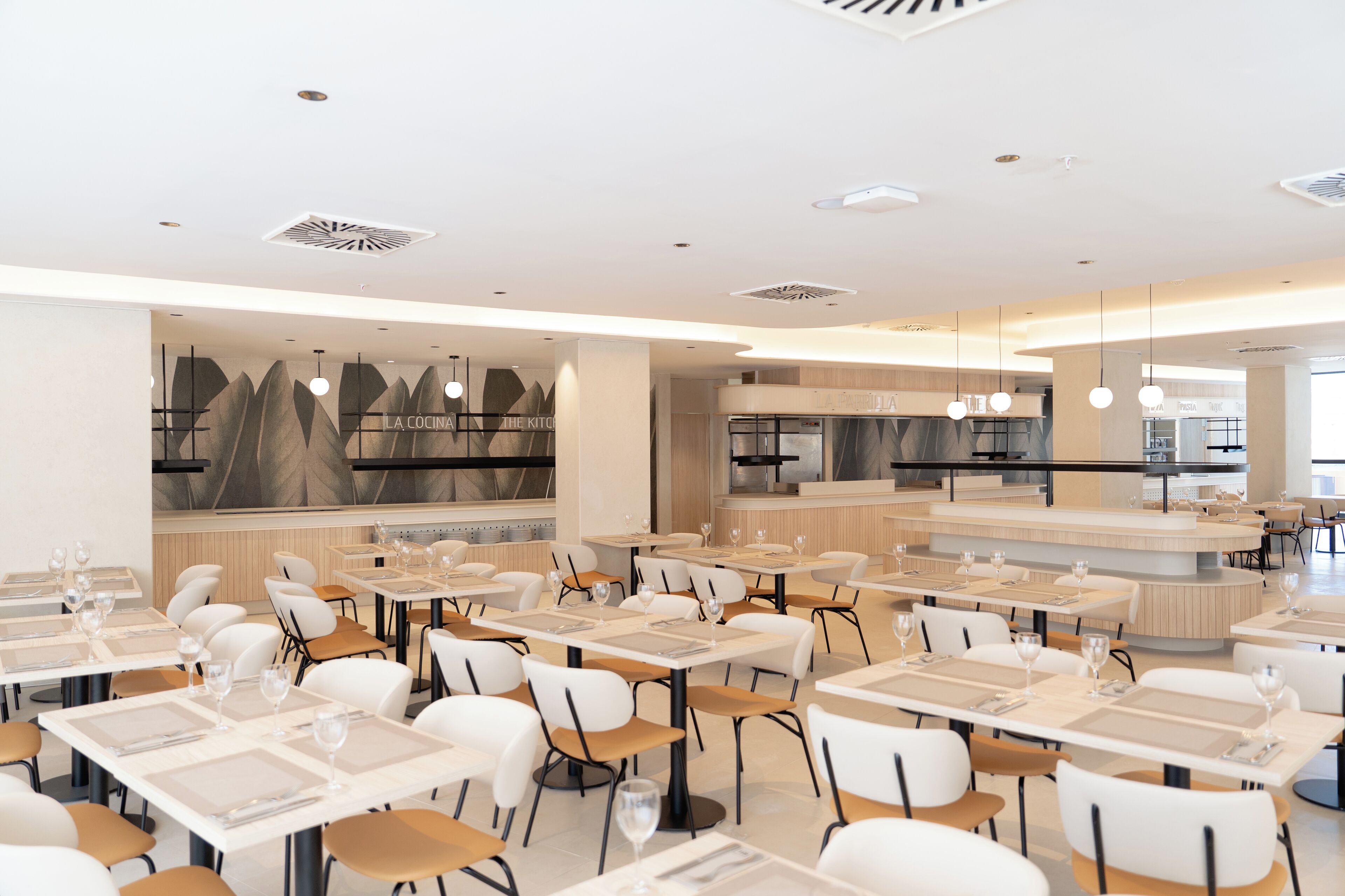 Un restaurant moderne doté de plusieurs tables et chaises, d’un espace cuisine et d’un éclairage suspendu.
