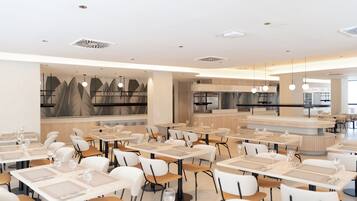 Un restaurante moderno con varias mesas y sillas, una zona de cocina e iluminación colgante.