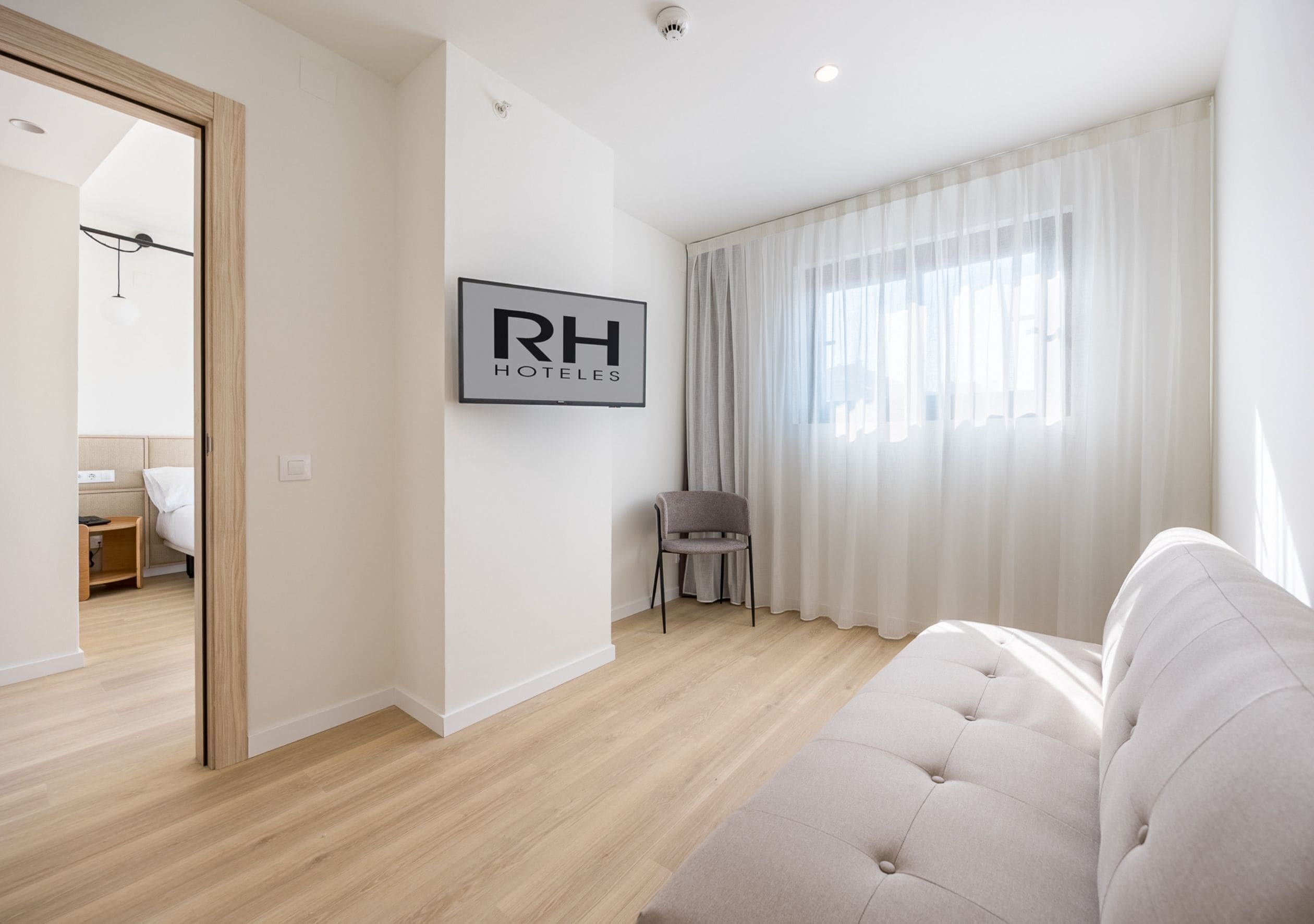 Una habitación de hotel moderna con un ventanal grande, un sofá, una silla y un cartel de RH HOTELS.