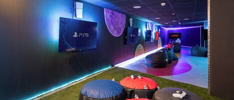 Un salón de juegos con sillones tipo bean bag, una vitrina con PlayStation 5 y un pasillo iluminado con luces de neón.