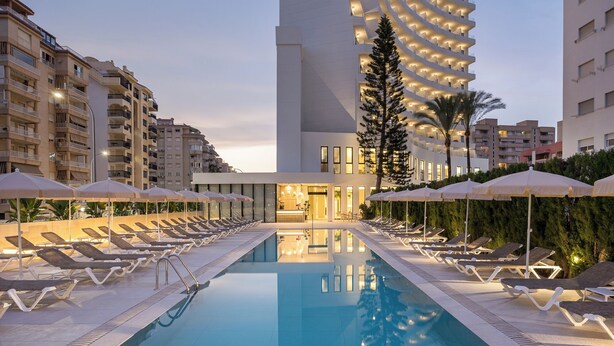 Una piscina con sillas de descanso y sombrillas, rodeada de edificios modernos.