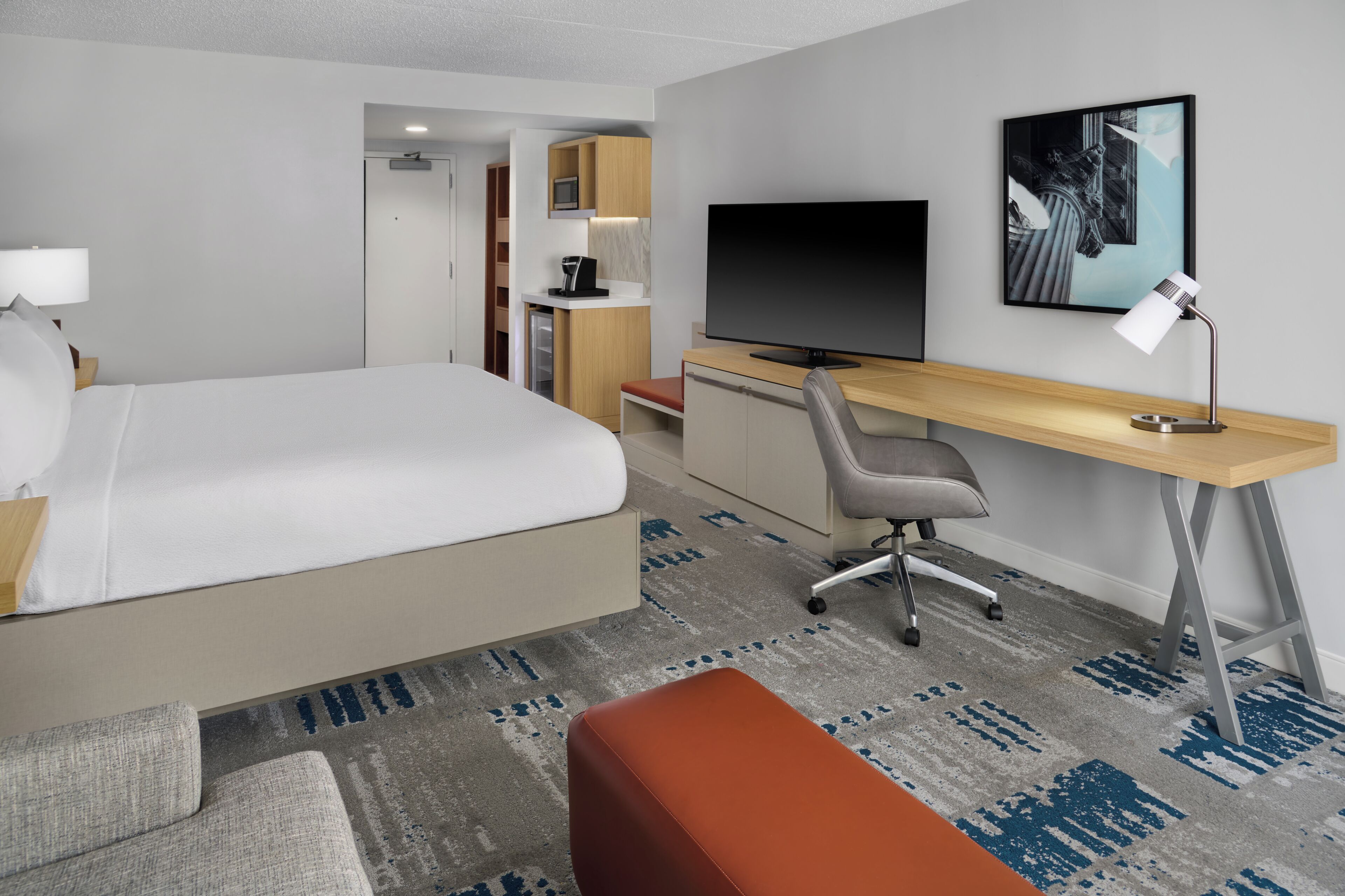 A hotel room with a bed, a desk with a computer, a TV, a coffee machine, and a painting on the wall.