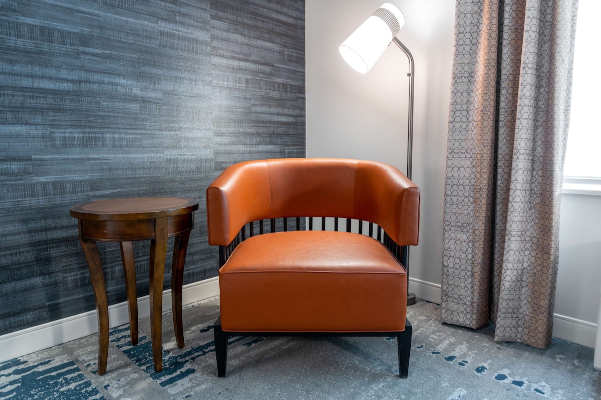 A room with a modern chair, a small wooden side table, a floor lamp, and patterned curtains.