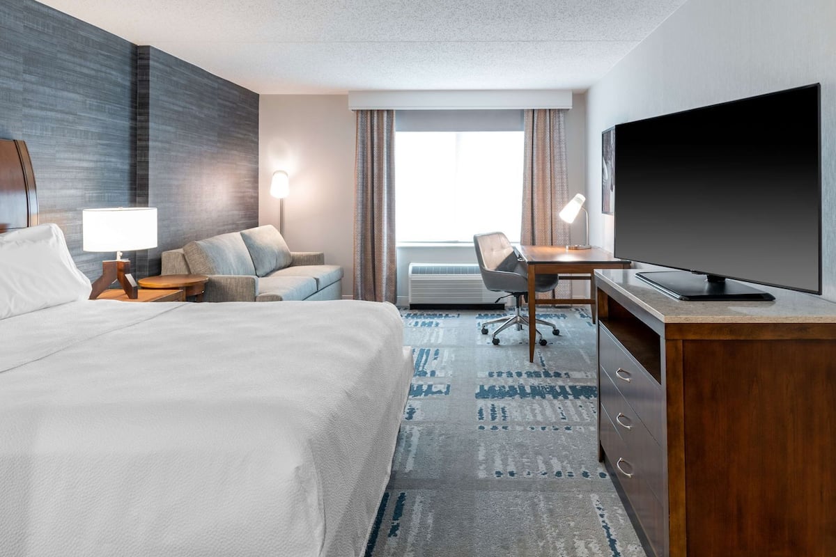 A hotel room with a bed, a desk with a TV, a chair, a lamp, and a window with curtains.
