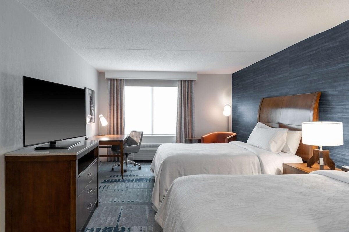 A hotel room with two beds, a desk with a chair, a television, and a large window with curtains.