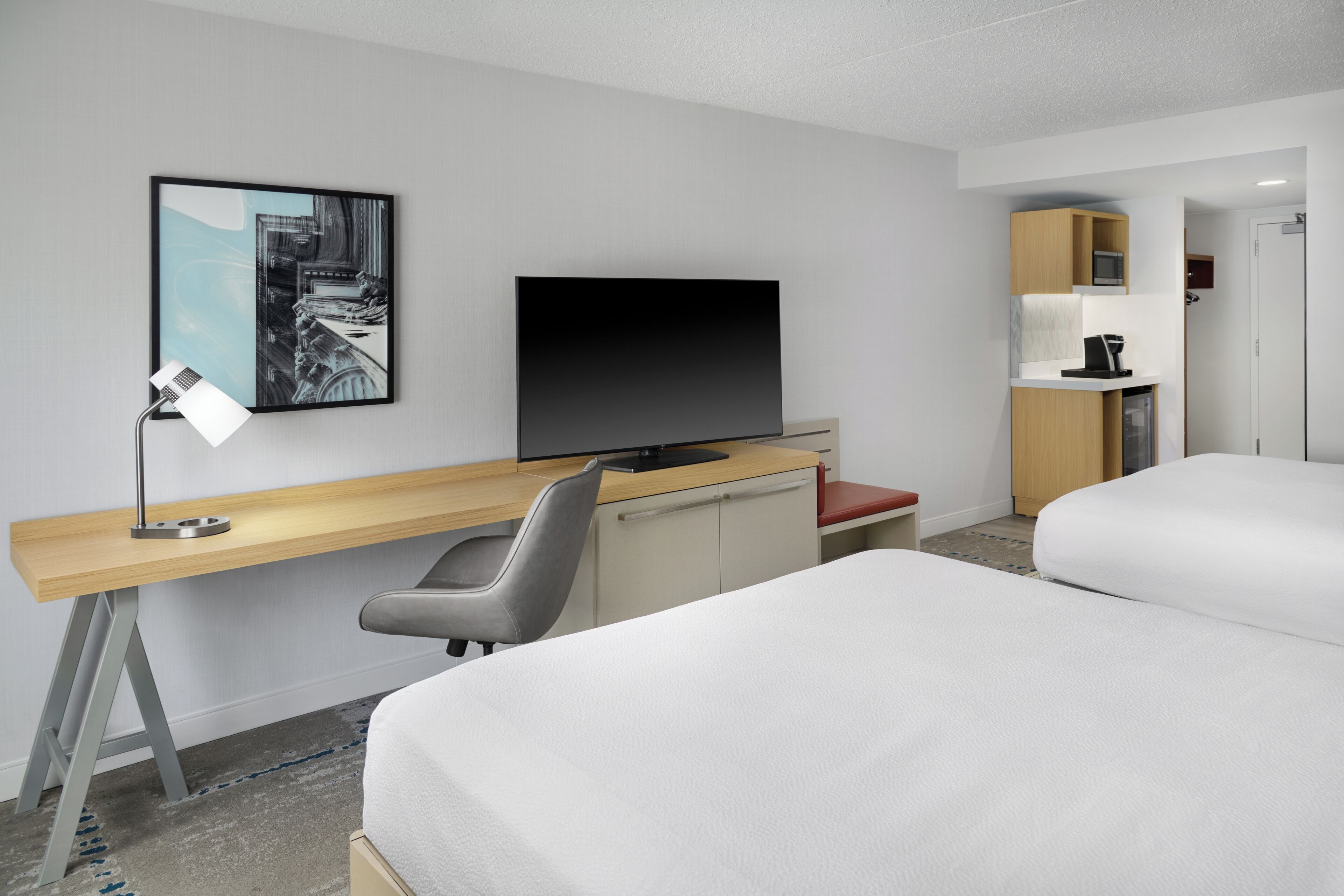 A hotel room with a bed, a desk with a computer, a TV, a chair, and a kitchenette.
