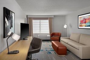 A hotel room with a desk, chair, sofa, TV, air conditioning unit, and artwork on the walls. - Hilton Garden Inn Arlington/Courthouse Plaza (Arlington)