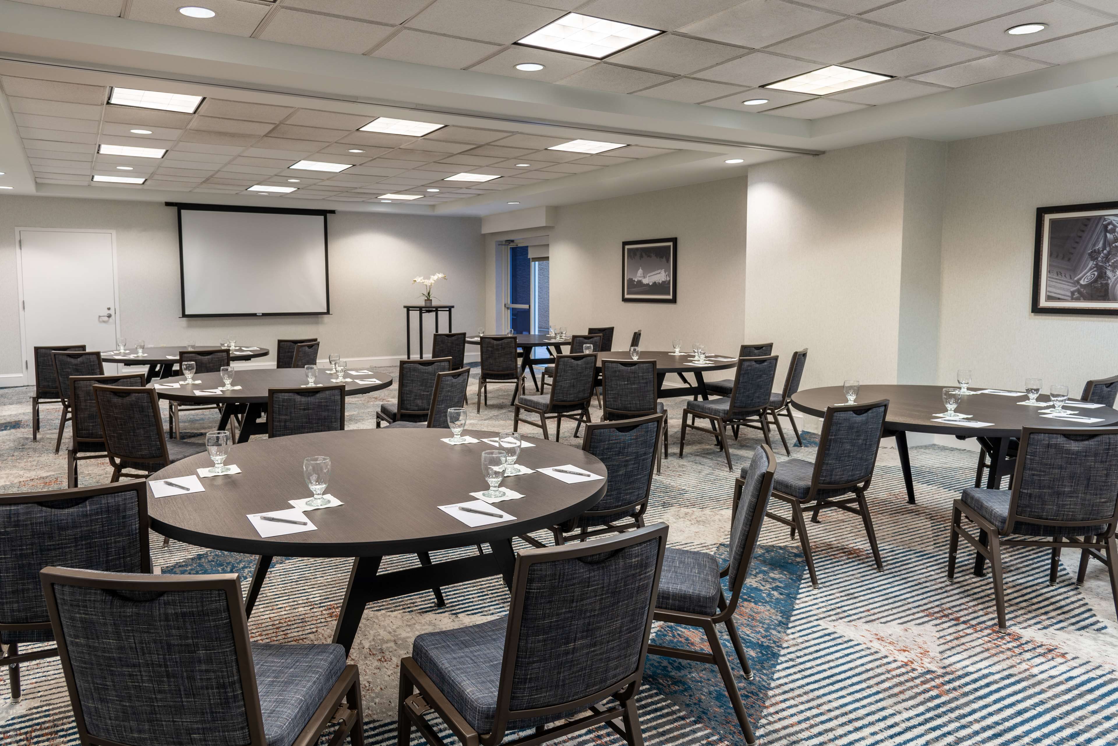 A conference room with multiple tables and chairs, a projector screen, and a carpeted floor.