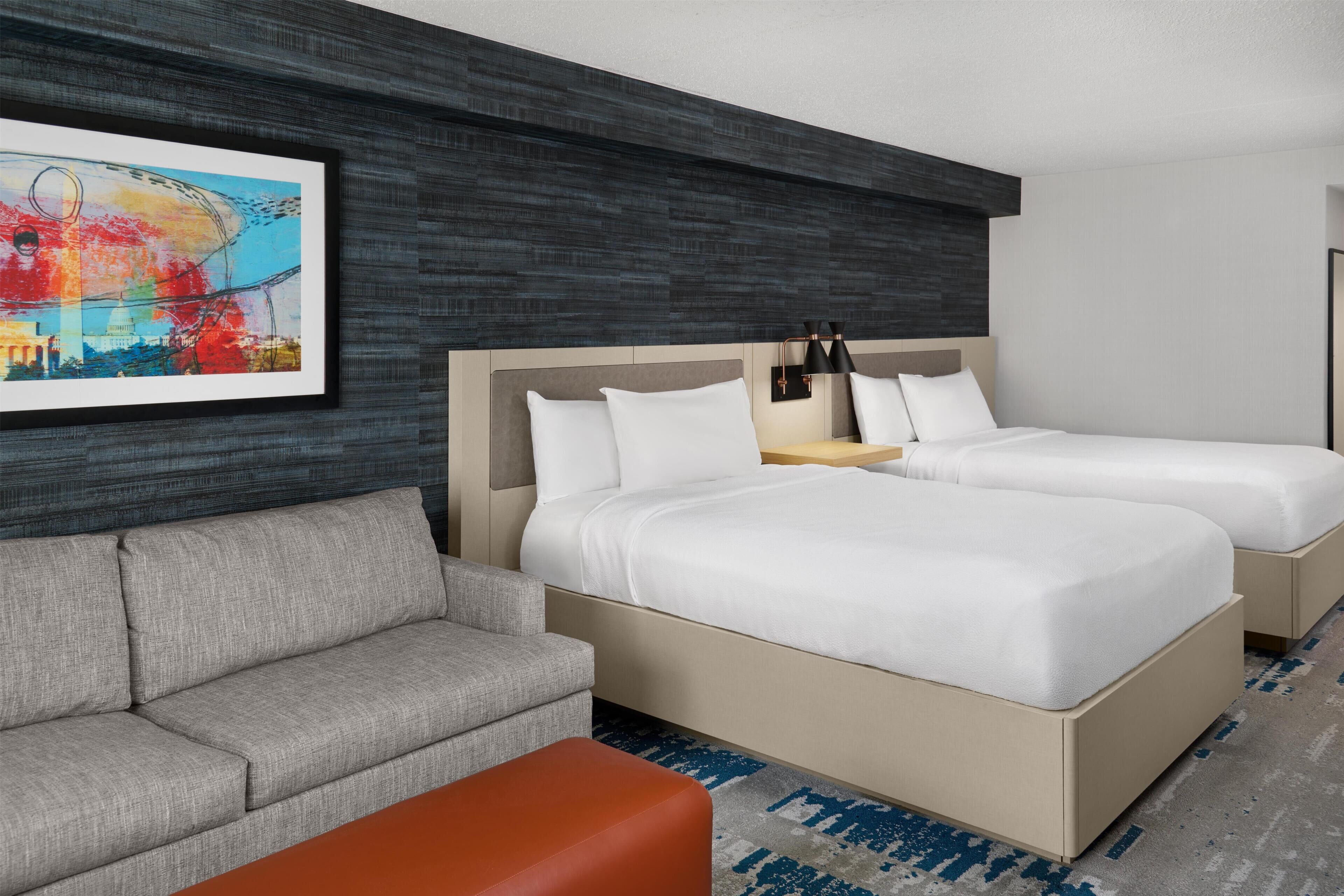 A hotel room with two beds, a gray sofa, an orange ottoman, and a framed artwork on the wall.