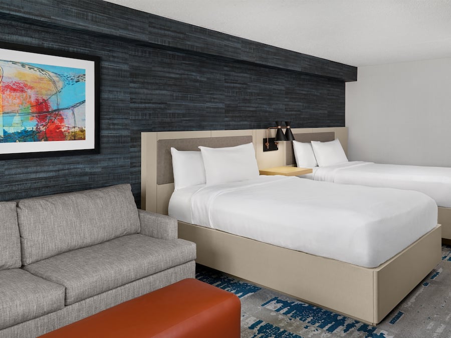 A hotel room with two beds, a gray sofa, an orange ottoman, and a framed artwork on the wall.