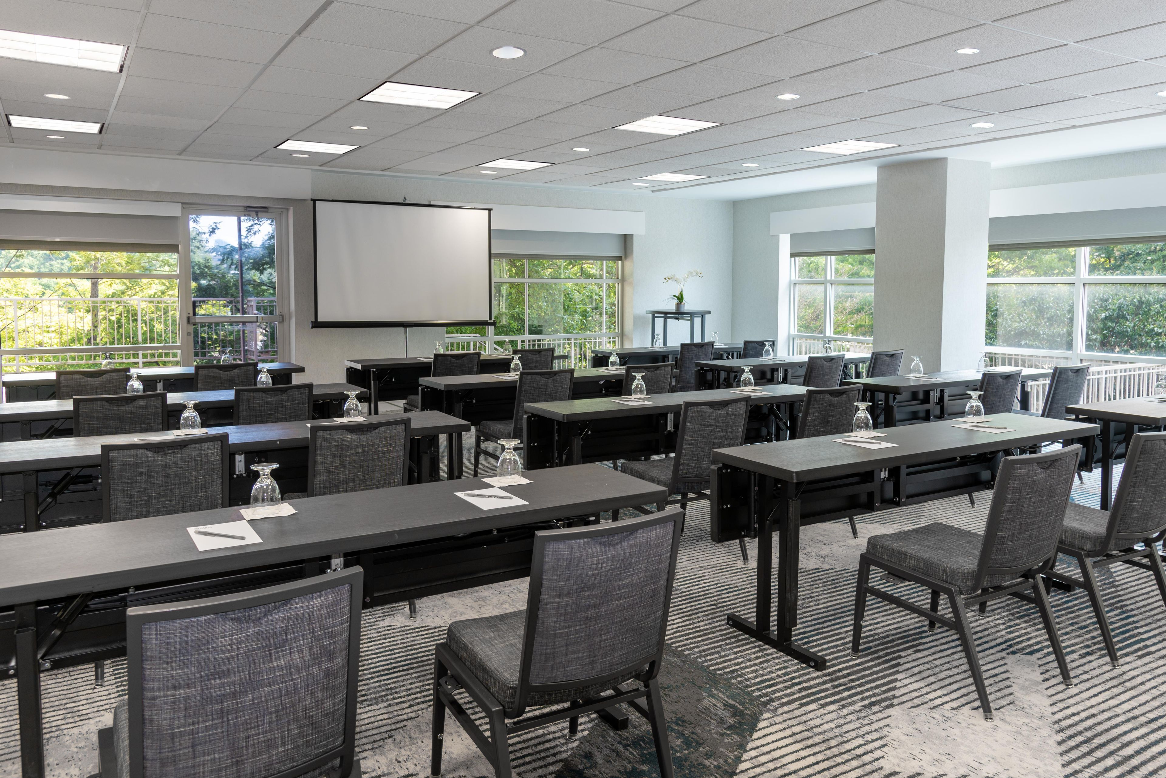 A conference room with multiple tables and chairs, a projector screen, and large windows with a view of greenery.