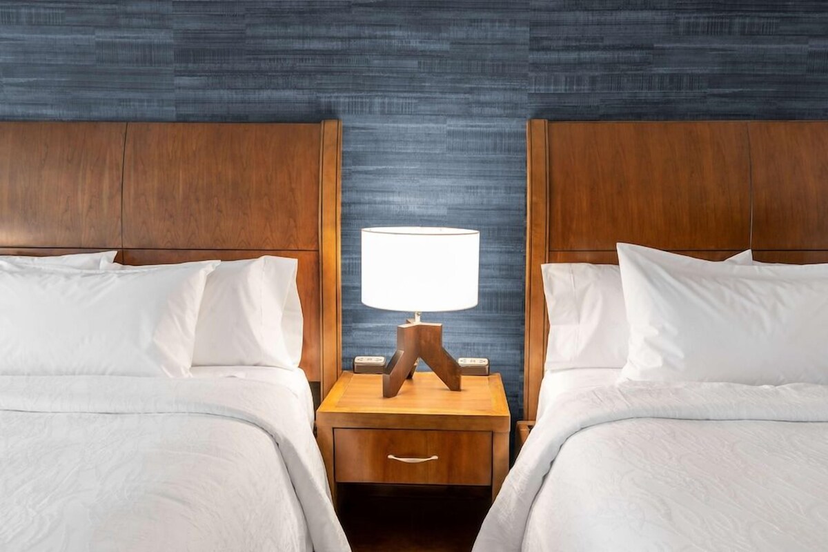 A hotel room with two beds, wooden headboards, a nightstand with a lamp, and a textured blue wall.