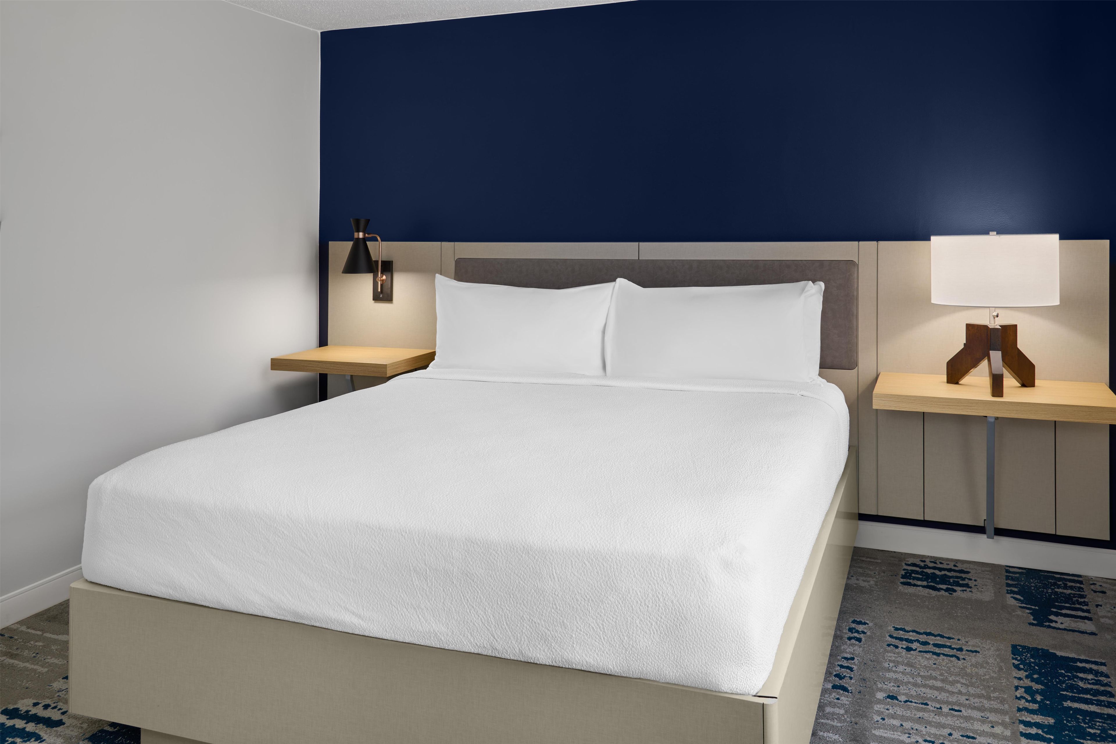 A hotel room with a bed, two bedside tables, a nightstand, and a wall-mounted lamp.