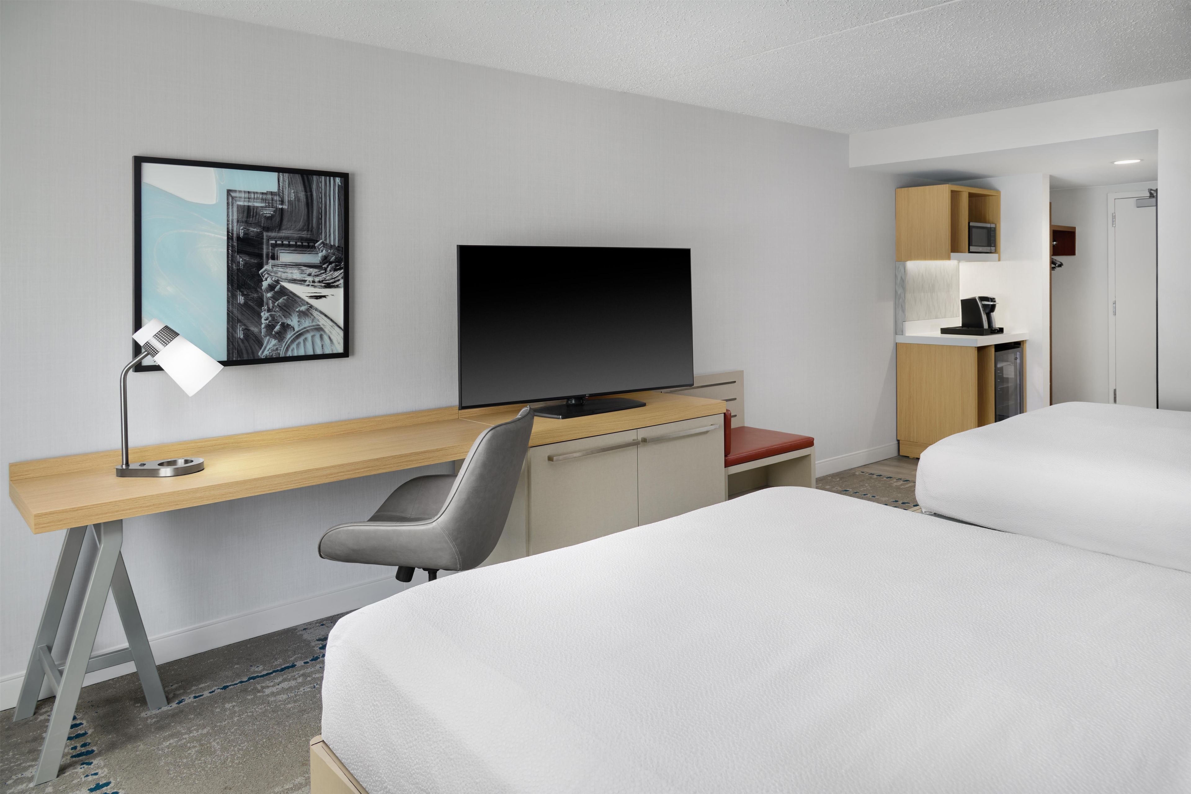A hotel room with a bed, a desk with a computer, a TV, a chair, and a small kitchenette.
