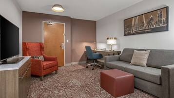 Quarto de hotel com televisão, uma poltrona vermelha, um sofá cinzento, uma secretária com abajur e um quadro com vista para a cidade.