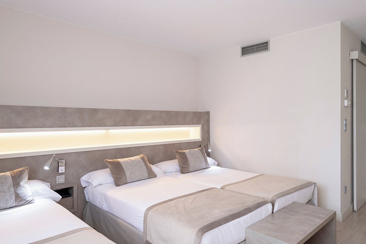 A hotel room with two beds, a bedside table, a lamp, and a window with a view.