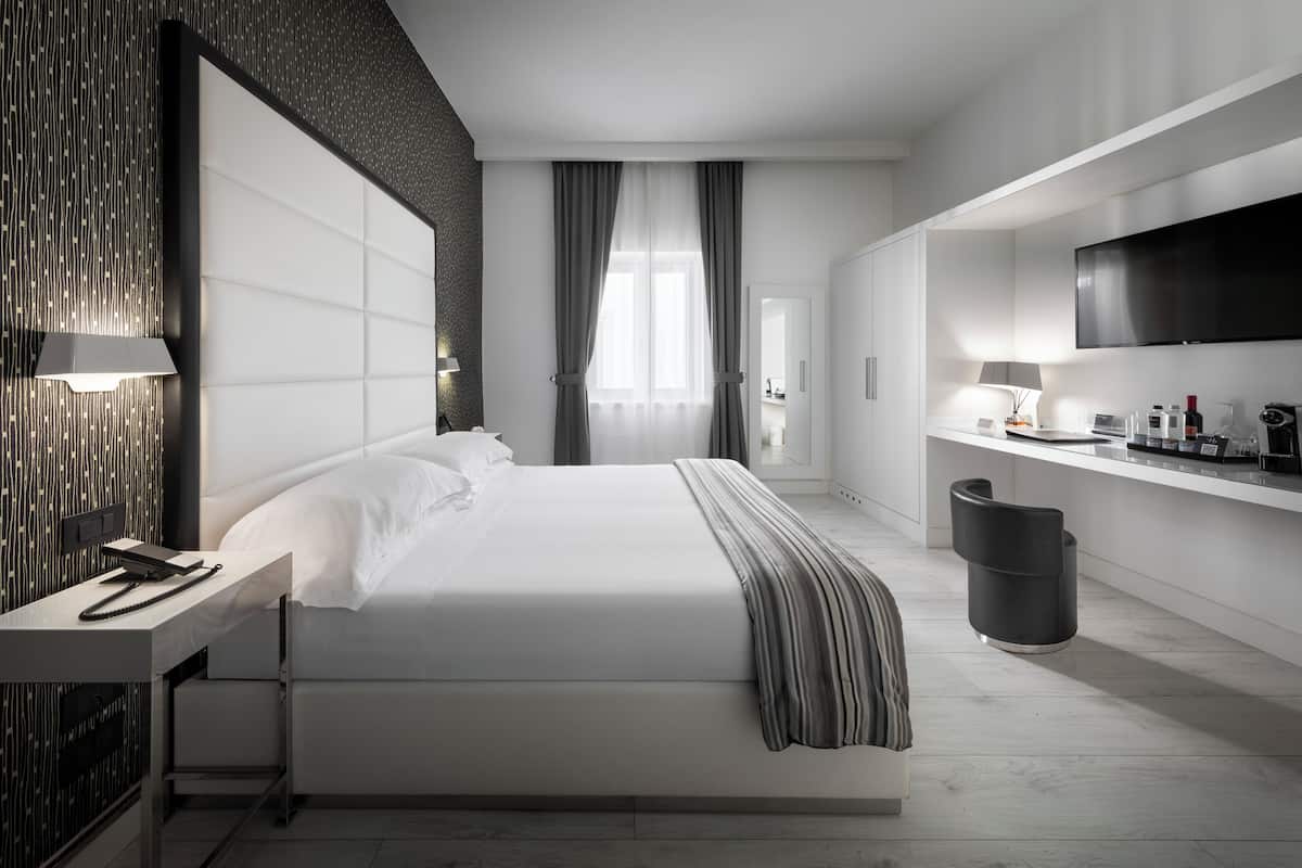 A modern hotel room with a large bed, a desk with a lamp, a TV, and a bathroom visible through a door.