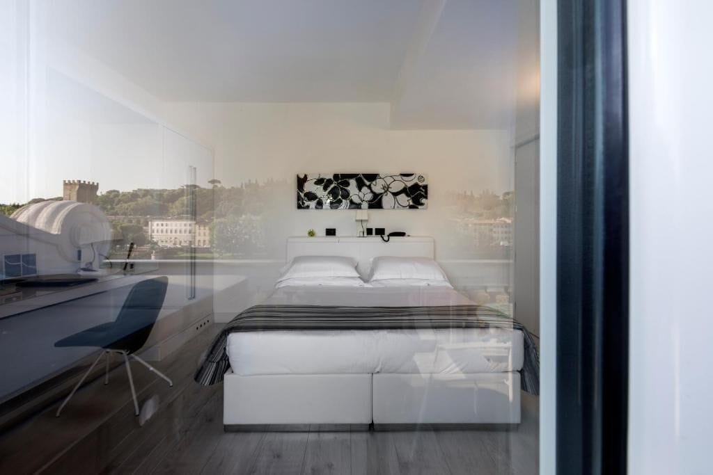 A modern bedroom with a large bed, a desk, and a chair. There is a city view through the window.