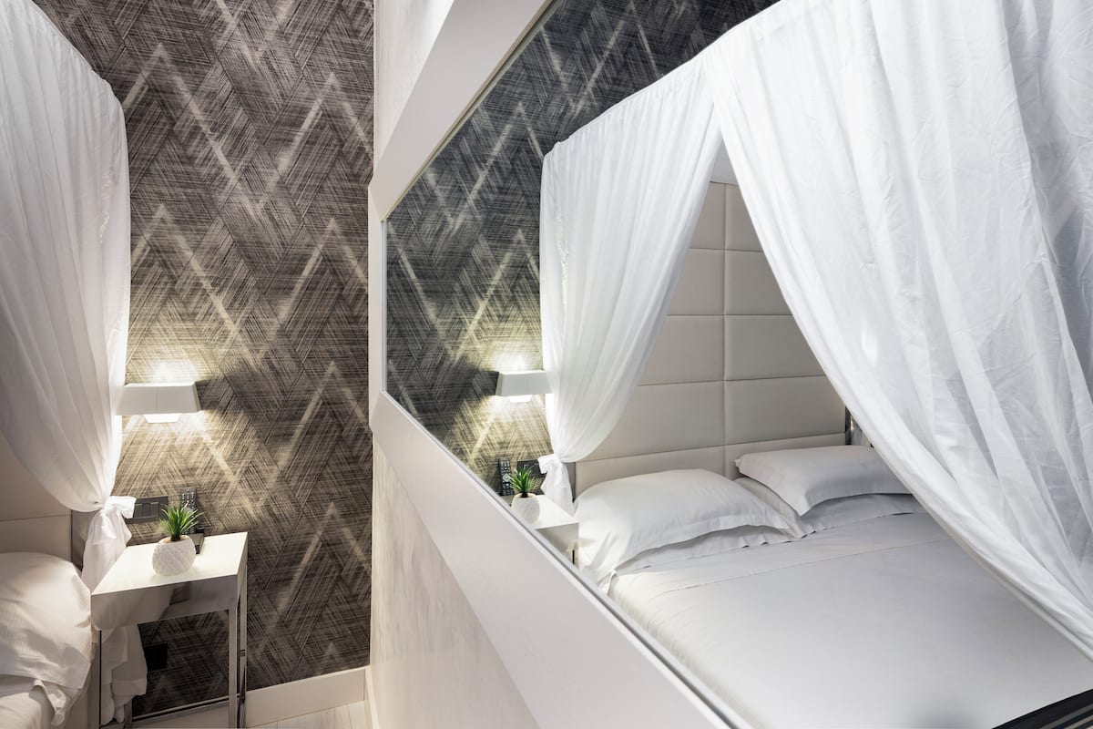 A modern bedroom with a geometric patterned wall, a white bed with a canopy, a bedside table with a lamp, and a small plant.