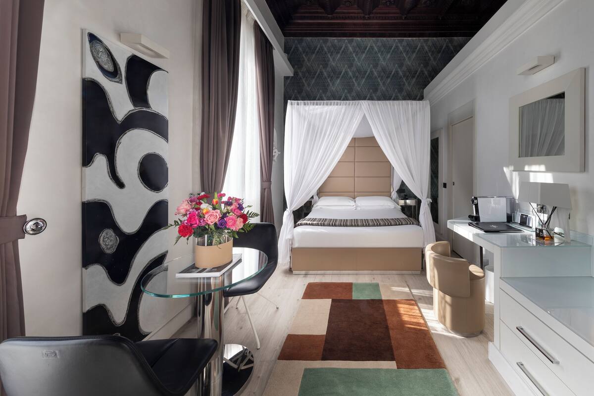 A modern hotel room with a bed, a desk, a chair, and a small table with flowers.
